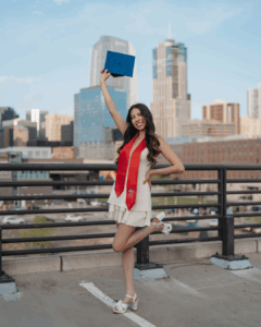 A young woman wearing a white dress and red graduation stole stands on a rooftop parking lot, holding a blue graduation cap high in the air with a bright smile. The city skyline, with tall modern buildings and a clear blue sky, provides a celebratory urban backdrop.