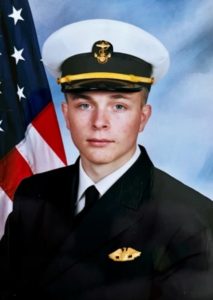 A front facing photo of a white man in military wear with an American flag behind him.